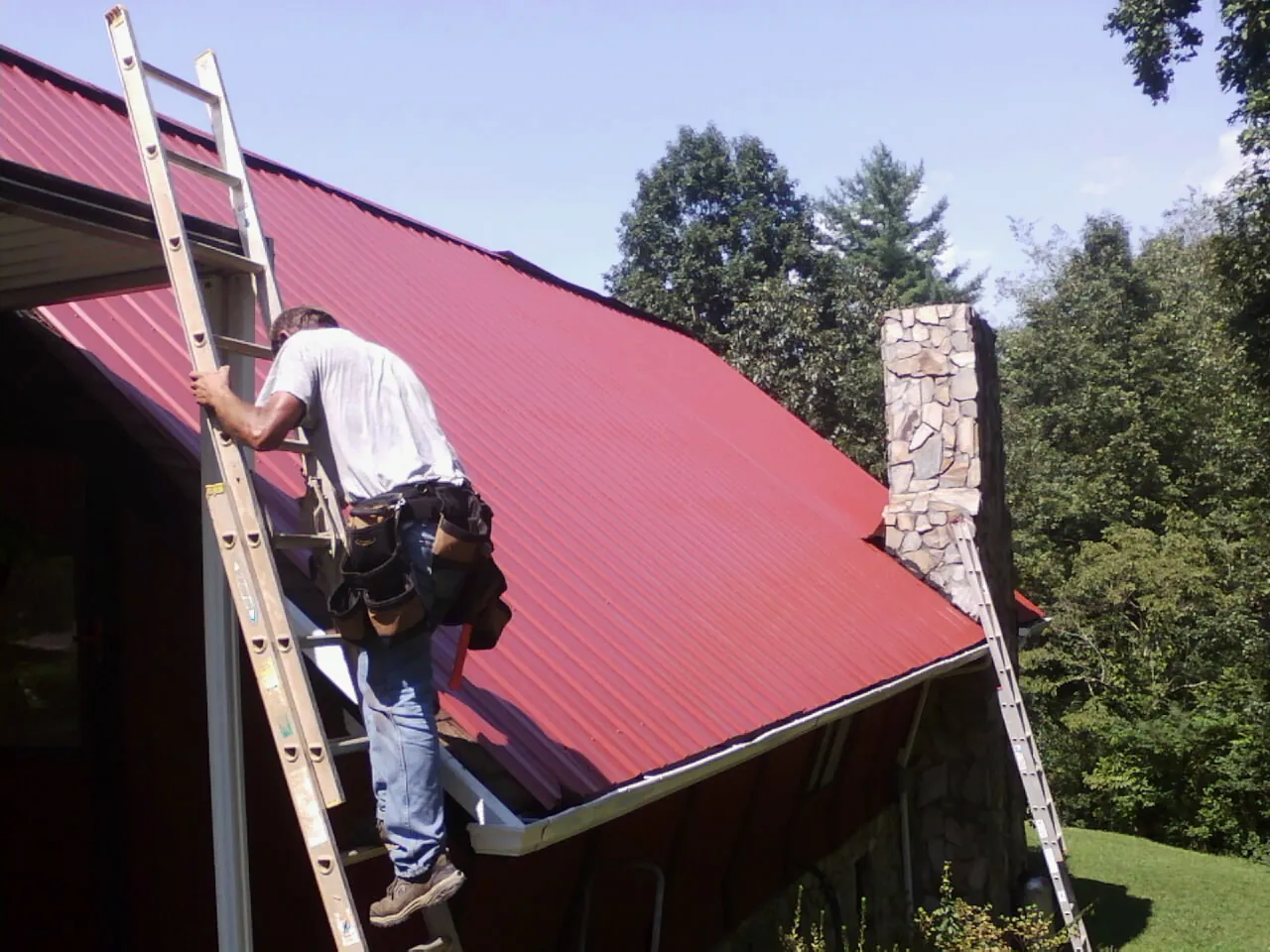 Roofer on a ladder beside a red metal roof and stone chimney — Soffit & Fascia Repair by Roofing Brothers in Sewell